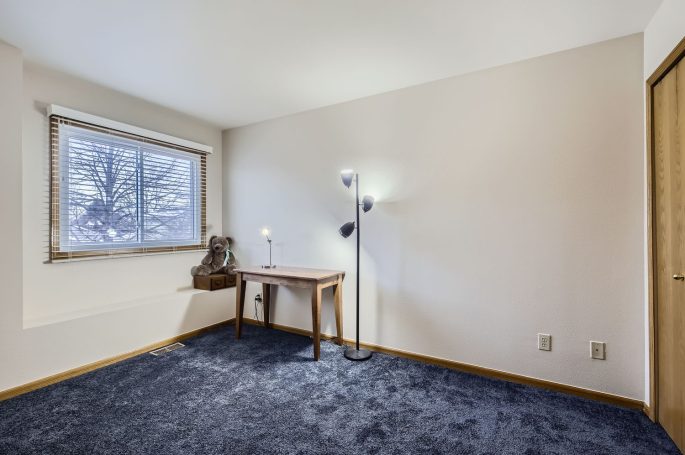 A vacant room with blue carpet, a wooden table, a lamp, and a window.